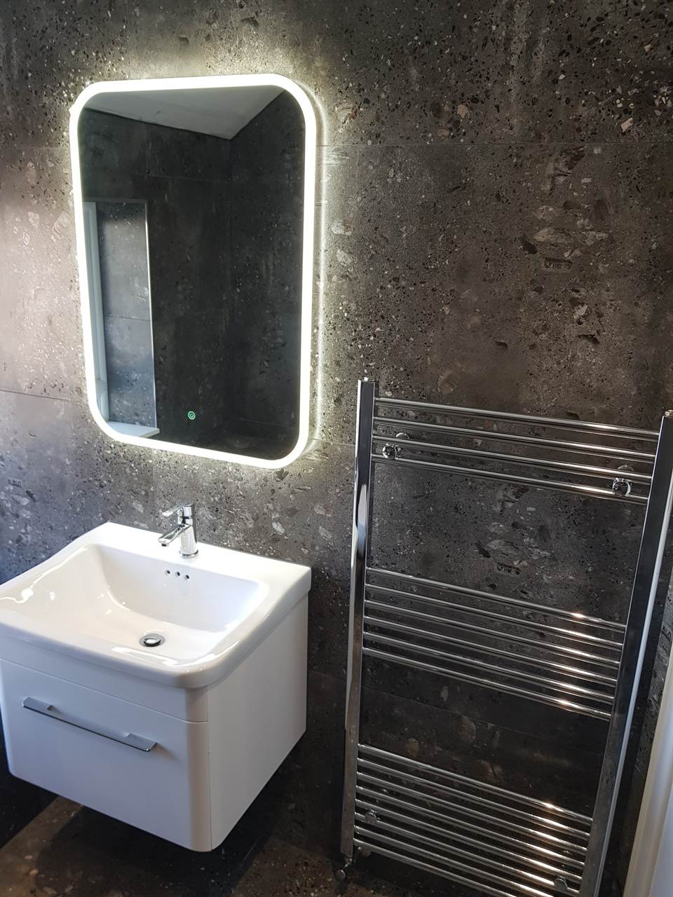 Bathroom with a backlit mirror, white wall-hung basin and chrome towel radiator against charcoal terrazzo-effect tiles